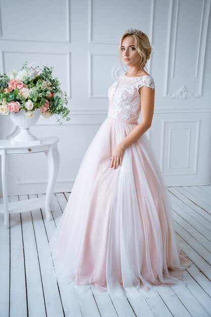 Una bella sposa con i capelli e il trucco si trova in un delicato abito da sposa rosa in un arredamento leggero con fiori