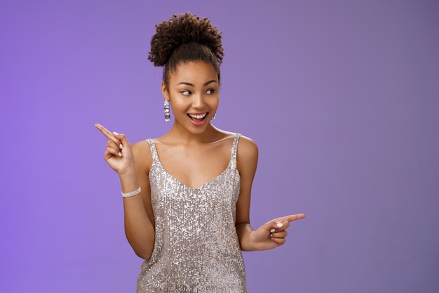 Una bella studentessa afro-americana glamour eccitata partecipa a un evento di lusso in argento lucido vestito perfetto ballare guardarsi intorno indicando destra sinistra sorridendo varianti compiaciute fare una scelta.