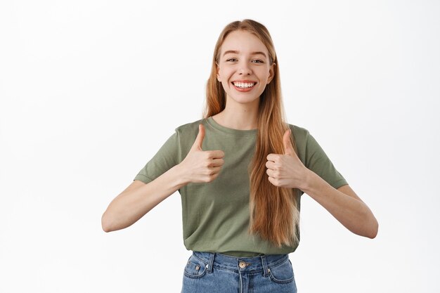 Una bella studentessa sorridente mostra i pollici in su, sembra soddisfatta e annuisce in segno di approvazione, darle supporto, consigliare il prodotto, elogiare la qualità eccellente, in piedi sul muro bianco