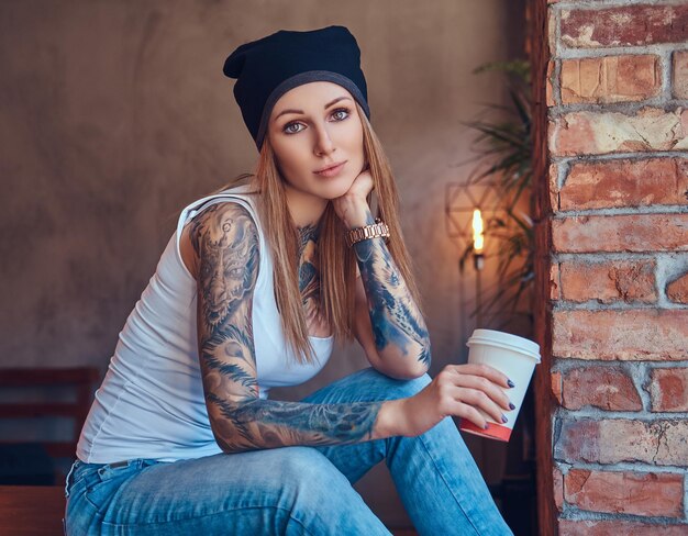 Una bionda sexy tatuata con una maglietta e un cappello con una tazza di caffè in una stanza con interni soppalcati.