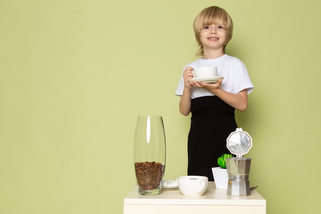 Una bionda sorridente del bambino di vista frontale adorabile in maglietta bianca che prepara la bevanda del caffè sullo spazio colorato pietra