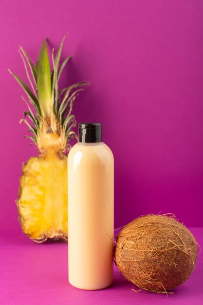 Una bottiglia di shampoo di plastica color crema vista frontale con tappo nero insieme a limoni ananas e cocco isolato su sfondo viola cosmetici bellezza frutti