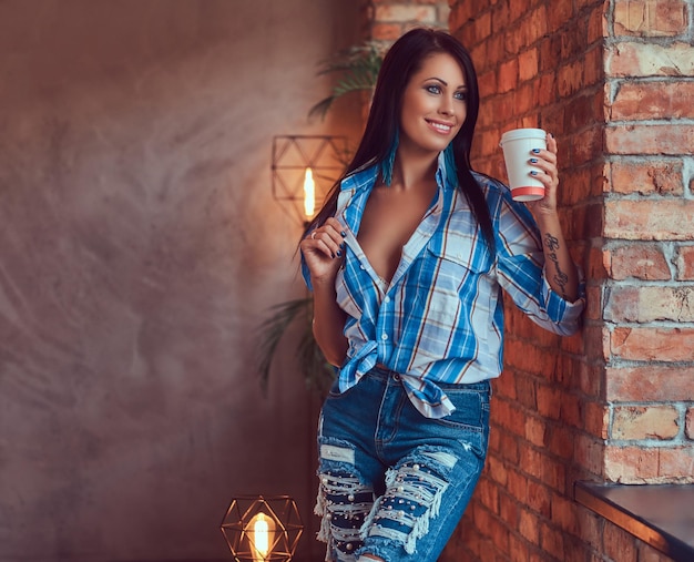 Una bruna sexy in una camicia di flanella e jeans tiene una tazza di caffè in posa contro un muro di mattoni.