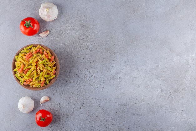 Una ciotola di argilla di pasta a spirale cruda multicolore con aglio e pomodori rossi freschi.