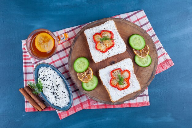 Una ciotola di formaggio, un bicchiere di tè accanto al pane al formaggio, fette di limone e cetriolo su una tavola, sul blu.