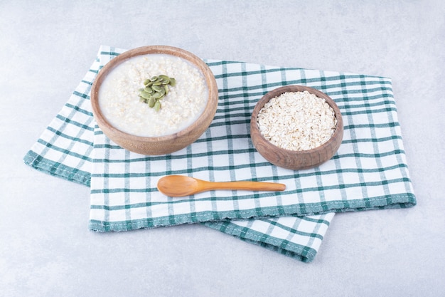 Una ciotola piena di avena cruda e una ciotola di farina d'avena condita con pepitas, accanto a un cucchiaio su un asciugamano sulla superficie di marmo