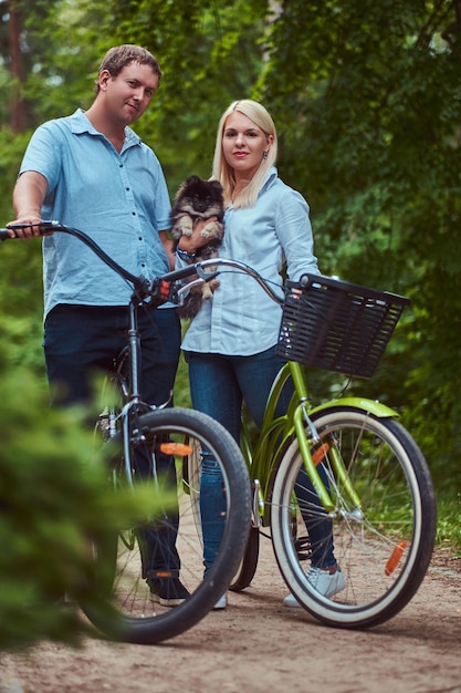 Una coppia attraente di una donna bionda e di un uomo vestito con abiti casual durante un giro in bicicletta con il loro piccolo spitz carino.