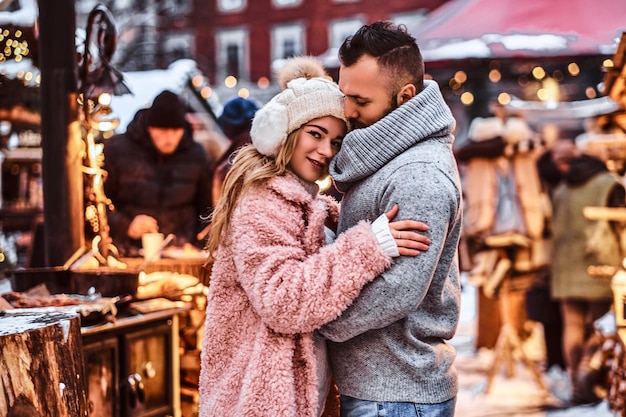 Una coppia attraente innamorata, un bell'uomo e una ragazza affascinante che si coccolano e si divertono a passare del tempo insieme mentre si trovano in piedi alla fiera invernale nel periodo natalizio