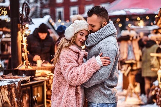 Una coppia attraente innamorata, un bell'uomo e una ragazza affascinante che si coccolano e si divertono a passare del tempo insieme mentre si trovano in piedi alla fiera invernale nel periodo natalizio