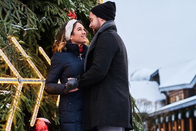 Una coppia felice che indossa abiti caldi si tiene per mano e si guarda l'un l'altro, in piedi vicino a un albero di Natale della città, godendosi il tempo trascorso insieme.