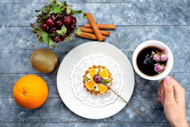 Una deliziosa piccola torta vista dall'alto con panna e frutta fresca a fette insieme a tè alla cannella sul biscotto alla frutta grigio-blu