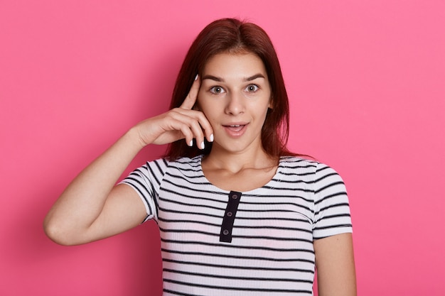 Una donna adorabile tiene l'indice sulla tempia, cerca di riunirsi con i pensieri, vestita con una maglietta a righe casual, isolata sul muro di rose, ha un'idea, sembra eccitata.