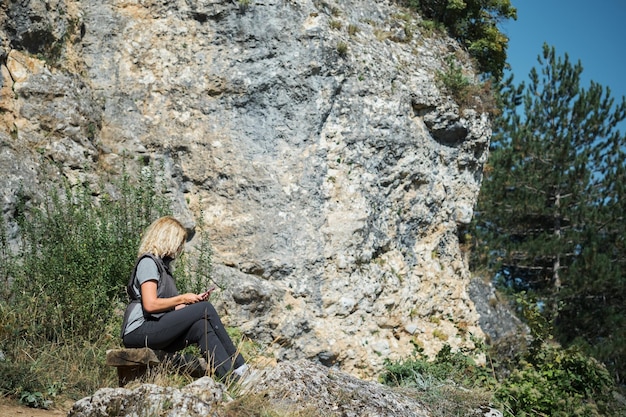 Una donna adulta si siede su una panchina sullo sfondo della roccia e guarda le vacanze sullo smartphone fuori città