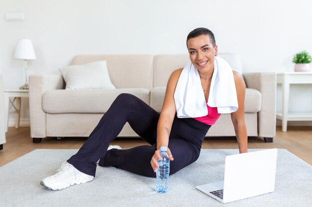 Una donna africana adulta fa esercizi di yoga e allenamento per la forza su un tappetino nel suo soggiorno. Segue un video di un corso di esercizi online sul suo laptop