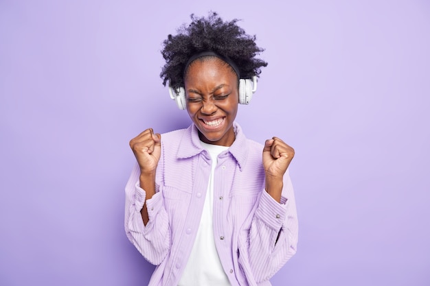 Una donna afroamericana di successo positiva stringe i pugni dalla gioia ascolta una nuova playlist musicale tramite cuffie wireless