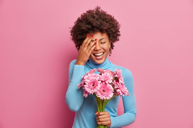 Una donna afroamericana felicissima ride ad alta voce, tiene il palmo sul viso, sta al chiuso con un mazzo di fiori di gerbera