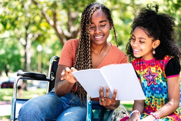 Una donna afroamericana su una sedia a rotelle si gode una giornata al parco con sua figlia mentre legge un libro insieme.