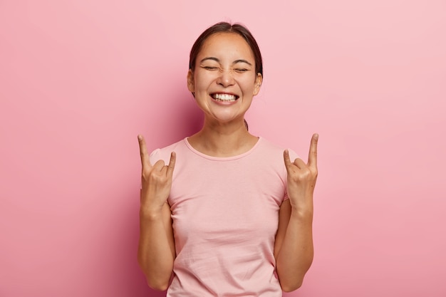 Una donna allegra dall'aspetto piacevole fa segni rock, gesticola con entrambe le mani, si sente ribelle, mostra heavy metal, si scatena, ascolta musica ad alto volume a casa, vestita in modo casual. La ragazza asiatica si diverte alla festa