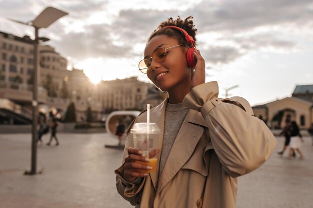 Una donna allegra in trench beige e occhiali da vista ama ascoltare la musica in cuffia fuori La signora beve il succo durante il tramonto