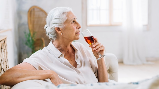 Una donna anziana che si siede sul sofà che tiene il vetro di alcool che daydreaming