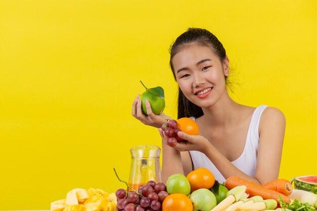 Una donna asiatica che indossa una canotta bianca. Entrambe le mani tengono la frutta e il tavolo è pieno di vari frutti.