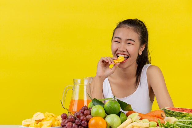 Una donna asiatica che indossa una canotta bianca. Mangiare ananas E il tavolo è pieno di vari tipi di frutta.