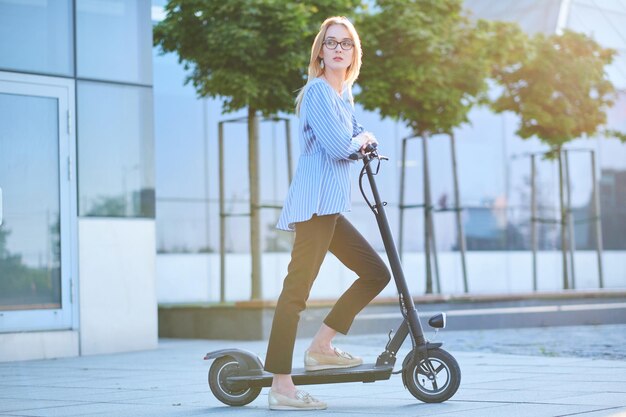 Una donna bionda e pensierosa con gli occhiali sta guidando il suo nuovo scooter elettrico per strada in una luminosa giornata di sole.