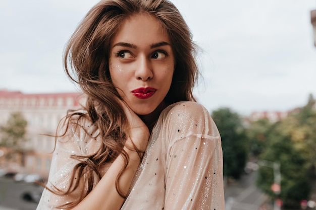 Una donna bruna abbronzata in camicia bianca posa di buon umore Una ragazza piuttosto allegra con le labbra rosse posa all'esterno con un sorriso