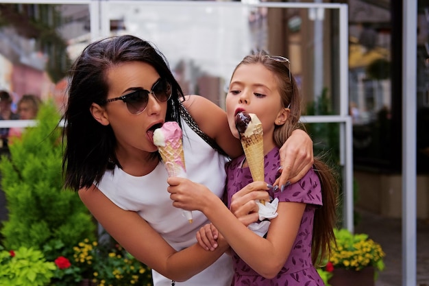 Una donna bruna attraente vestita con un abito bianco e occhiali da sole e una ragazza adolescente carina che mangia il gelato in una strada.