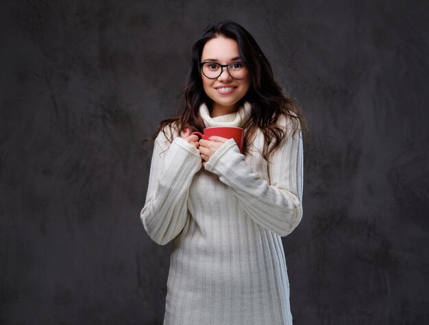 Una donna bruna emotiva e sorridente vestita con un caldo maglione a collo alto tiene una grande tazza di caffè rossa su sfondo grigio.