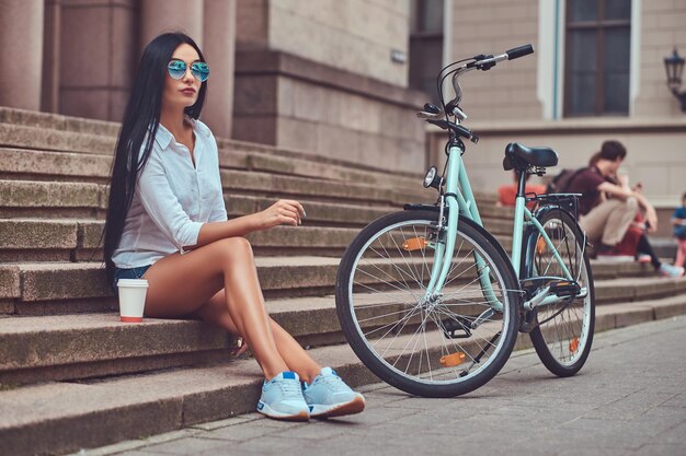 Una donna bruna sexy che indossa una camicetta e pantaloncini di jeans in occhiali da sole, rilassante dopo aver guidato su una bicicletta, seduta con una tazza di caffè sui gradini di una città.