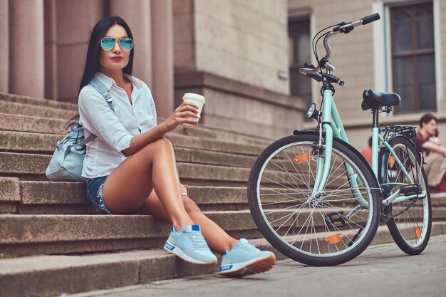 Una donna bruna sexy che indossa una camicetta e pantaloncini di jeans in occhiali da sole, rilassante dopo aver guidato su una bicicletta, seduta con una tazza di caffè sui gradini di una città.