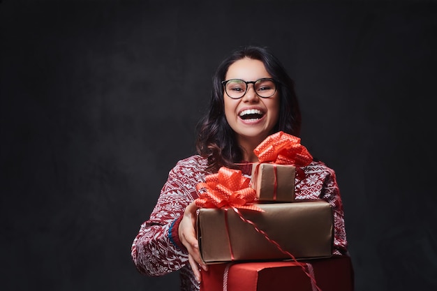 Una donna bruna sorridente vestita con un pullover rosso e occhiali da vista tiene i regali di Natale su sfondo grigio.