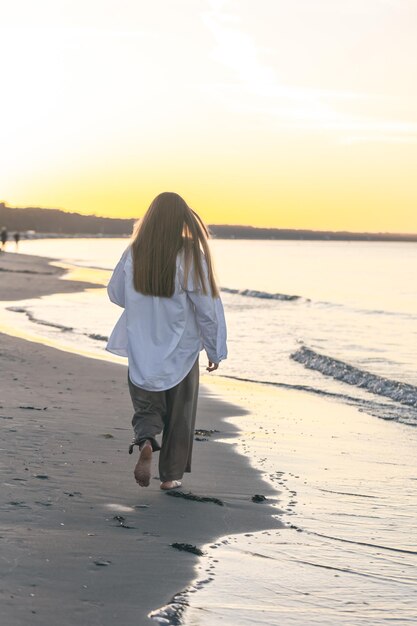 Una donna cammina lungo la spiaggia del mare al tramonto vista dal retro