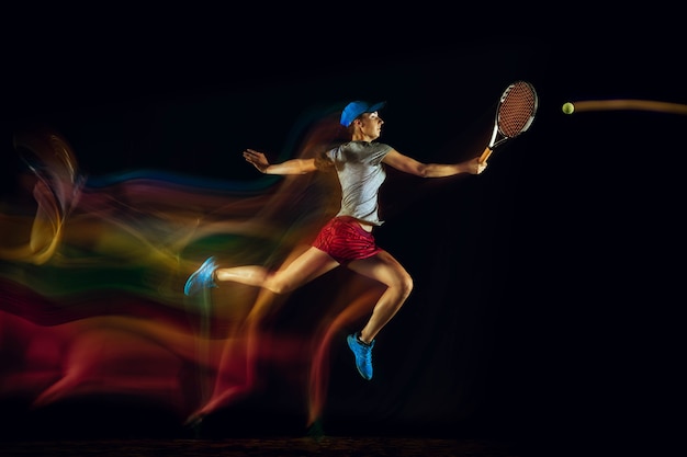 Una donna caucasica che gioca a tennis isolata sulla parete nera nella luce mista e stobe. Montare la giovane giocatrice in movimento o in azione durante il gioco sportivo. Concetto di movimento, sport, stile di vita sano.