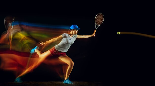 Una donna caucasica che gioca a tennis isolata sulla parete nera nella luce mista e stobe. Montare la giovane giocatrice in movimento o in azione durante il gioco sportivo. Concetto di movimento, sport, stile di vita sano.