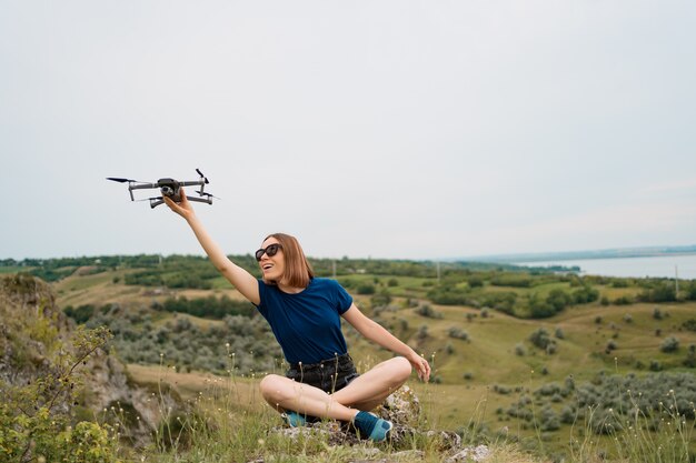 Una donna caucasica con un drone in mano, seduta su una verde collina rocciosa con cielo sullo sfondo