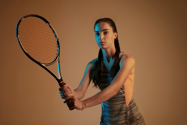 Una donna caucasica giocando a tennis isolato su sfondo marrone in luce mista e al neon. Montare la giovane giocatrice in movimento o in azione durante il gioco sportivo. Concetto di movimento, sport, stile di vita sano.