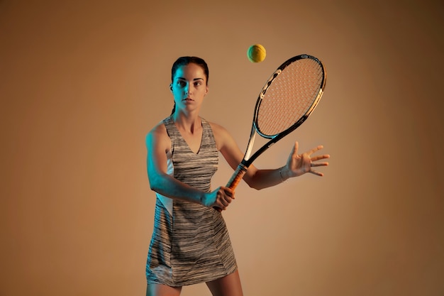 Una donna caucasica giocando a tennis isolato su sfondo marrone in luce mista e al neon. Montare la giovane giocatrice in movimento o in azione durante il gioco sportivo. Concetto di movimento, sport, stile di vita sano.