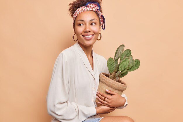 Una donna dai capelli ricci dalla pelle piuttosto scura si siede sembra felicemente lontana tiene una pentola di cactus succulenti spinosi