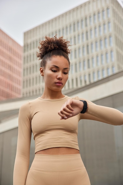 Una donna dai capelli ricci seri controlla i risultati dell'allenamento fitness con l'orologio da polso controlla le calorie bruciate dopo l'allenamento indossa il top corto e i leggings posa contro gli edifici urbani Stile di vita sportivo