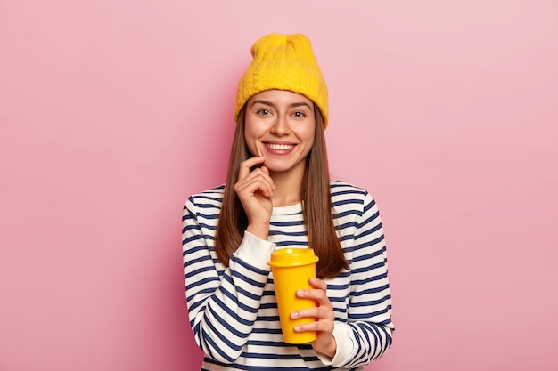 Una donna dall'aspetto piacevole adora il caffè, tiene la tazza da asporto, sorride piacevolmente, indossa un cappello giallo e un maglione a righe, posa sul muro rosa, gode dell'odore