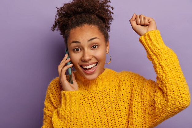 Una donna dalla pelle scura felicissima ha una conversazione mobile positiva, ascolta ottime notizie, alza il braccio e balla dalla gioia