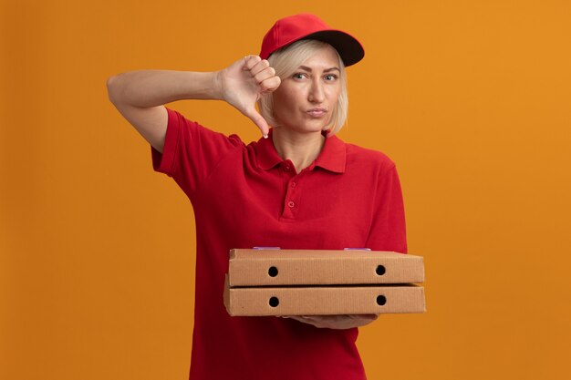 Una donna delle consegne bionda di mezza età scontenta in uniforme rossa e berretto che tiene in mano i pacchetti di pizza che mostra il pollice verso il basso