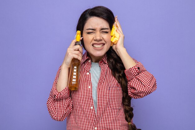 Una donna delle pulizie caucasica piuttosto spiacevole che tiene in mano panni per la pulizia e un detergente spray vicino alla sua testa