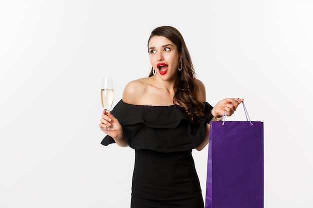 Una donna eccitata fa shopping e beve champagne, tiene in mano la borsa della spesa, sembra stupita, in piedi su sfondo bianco