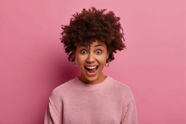 Una donna espressiva emotiva con l'acconciatura afro esclama e ha un'espressione stupita, tiene la bocca aperta, ha un'espressione gioiosa e divertente, vestita con un maglione rosa pastello, posa al coperto. Persone, eccitazione