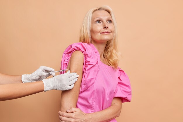 Una donna europea di mezza età bionda pensierosa riceve la vaccinazione a braccio contro il coronavirus indossa una camicetta rosa isolata su un muro marrone