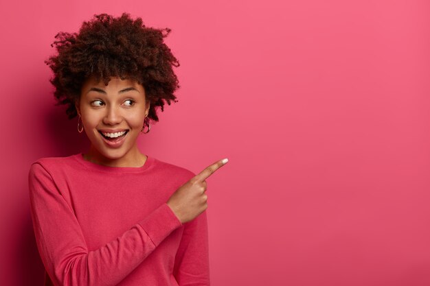 Una donna felice e positiva con taglio di capelli afro punta il dito indice nell'angolo in alto a destra, consiglia di provare il prodotto, indossa un maglione cremisi