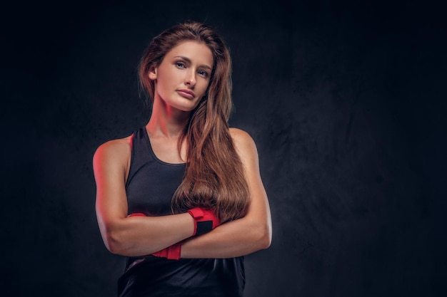 Una donna fisicamente in forma con i capelli lunghi è in piedi incrociando le mani su uno sfondo scuro.